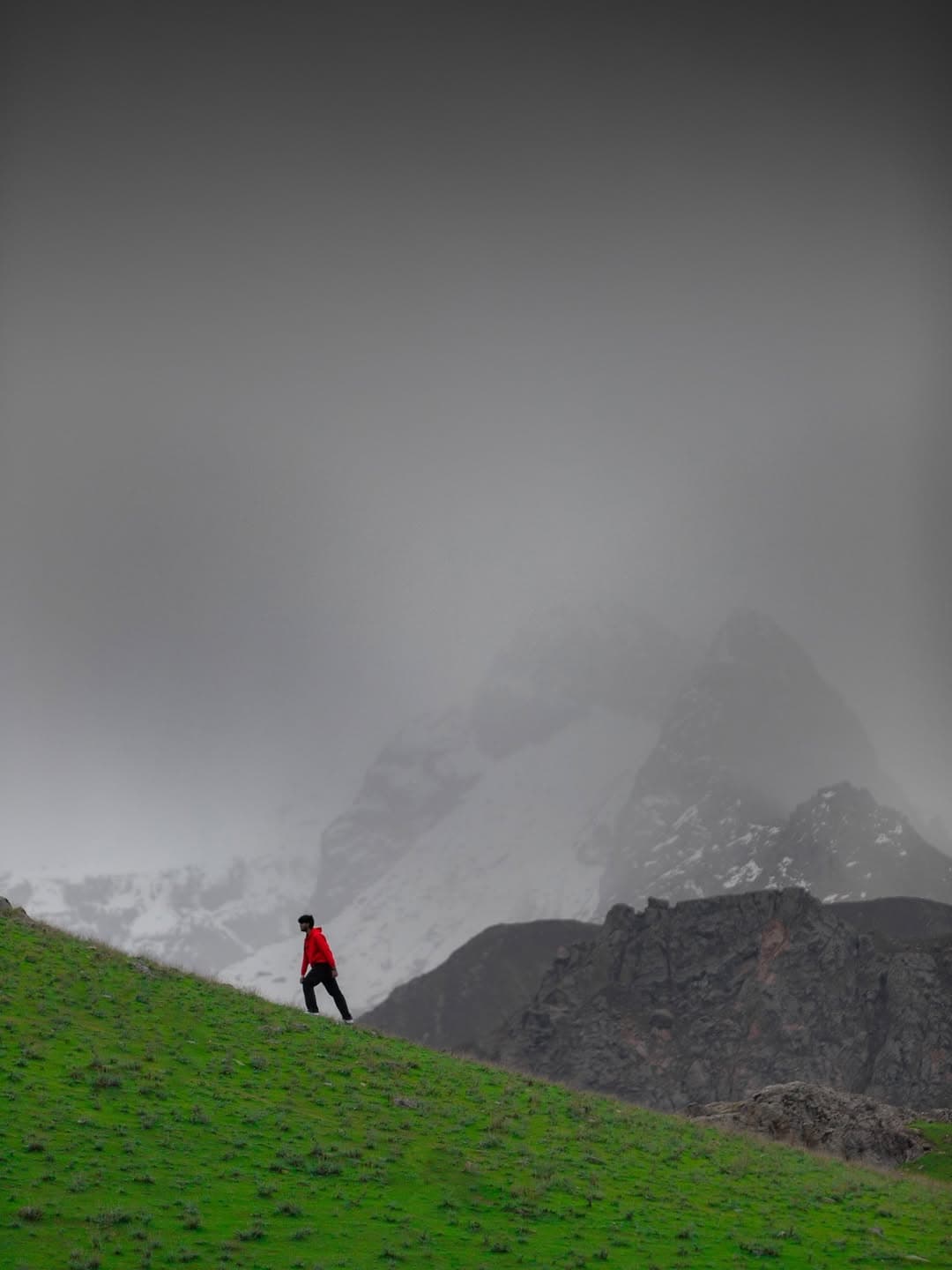 Lone hiker in red jacket on green slope with misty mountain peaks